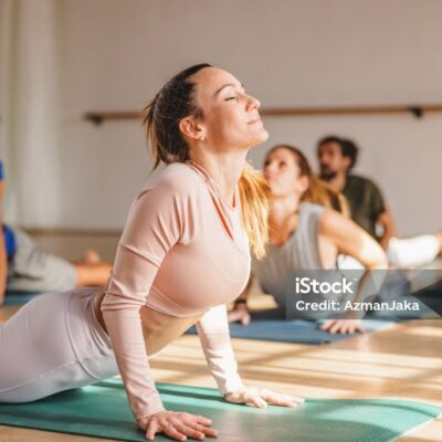 An adult caucasian female in a yoga class doing the cobra yoga pose with her eyes closed. She is lying on her stomach on a yoga mat. She is surrounded by other yoga practitioners. The studio is illuminated by sunrays.